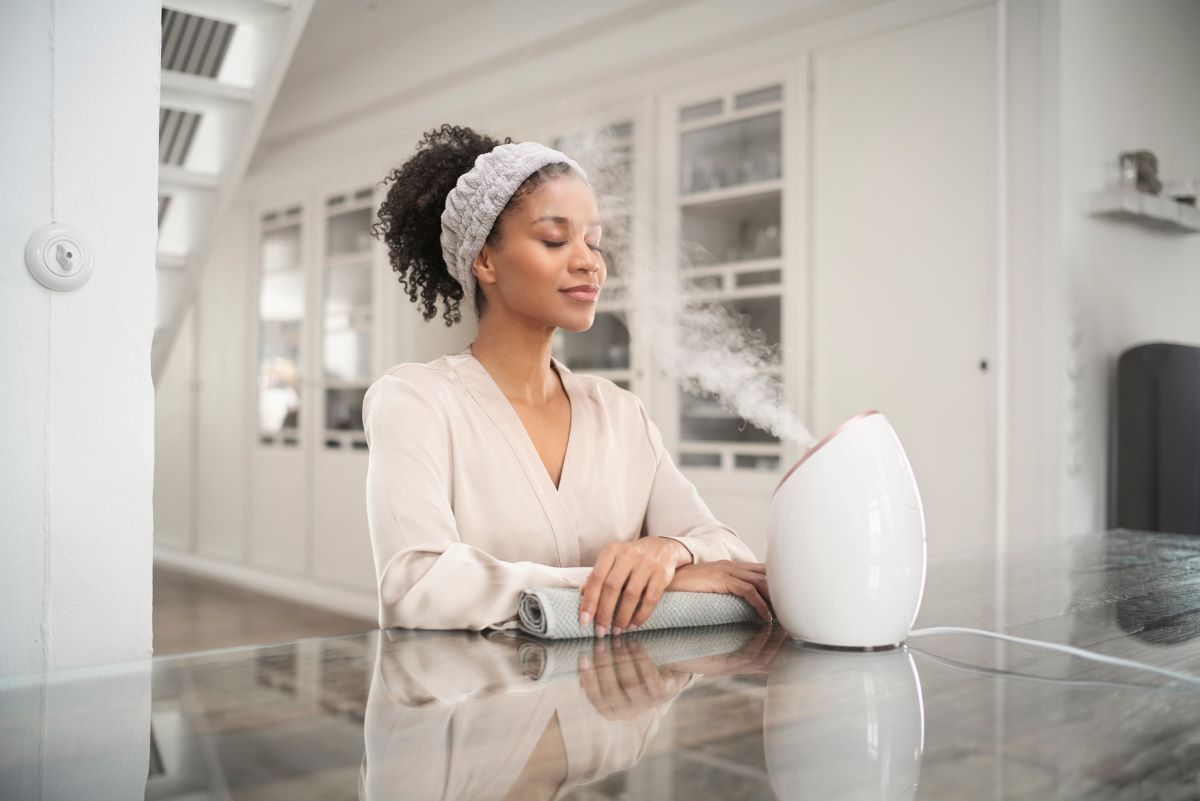 Woman sitting at a table, smiling while using a medisana facial sauna for skincare.